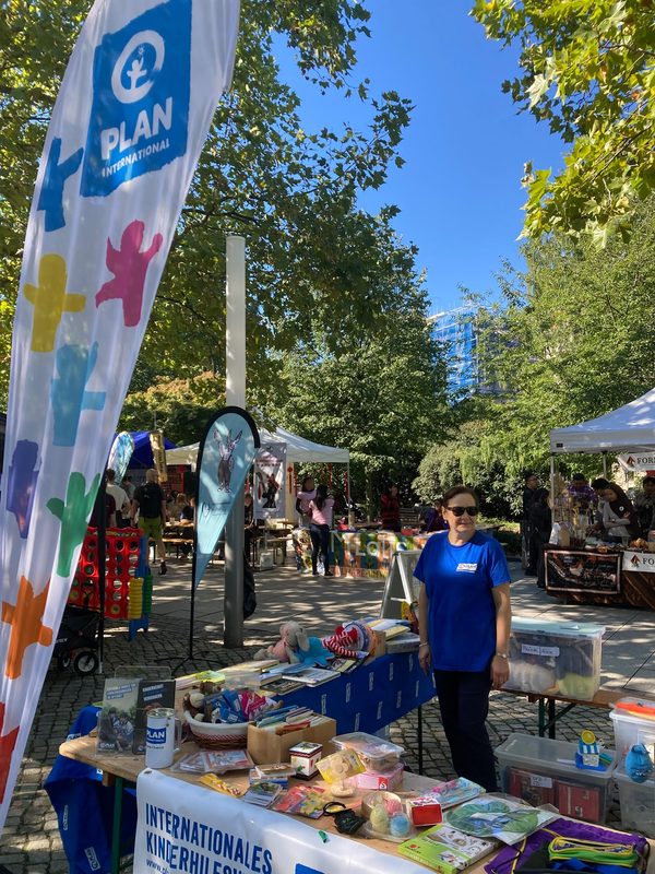 Auf einem Markt im Freien steht eine Frau hinter einem Tisch, der mit Büchern und Kleinigkeiten bedeckt ist; daneben ist ein Banner von Plan International zu sehen.